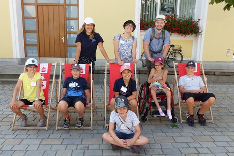 Das Foto zeigt die Teilnehmend und ihre Betreuerinnen und Betreuer der inklusiven Ferienfreizeit vor einem Haus. Die Teilnehmenden sitzen glücklich in Liegestühlen.