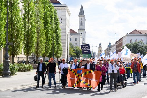 Demonstrierende Menschen mit Bannern und Fahnen auf einer Stra&szlig;e vor historischen Geb&auml;uden bei sonnigem Wetter