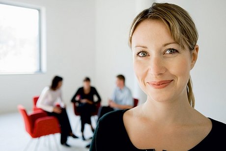 Porträtfoto einer Frau. Im Hintergrund sieht man drei Personen auf Stühlen sitzen.