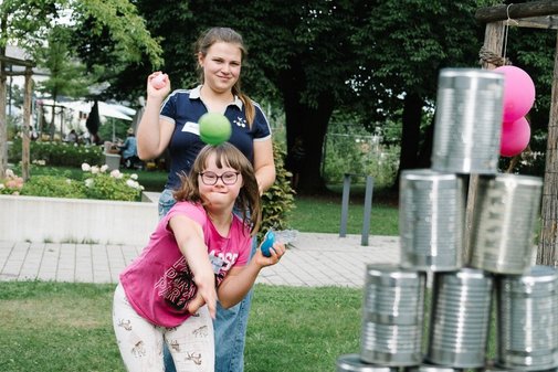 Zwei Freizeitteilnehmerinnen auf einer Wiese werfen Wasserbomben auf aufgestapelte Dosen. 