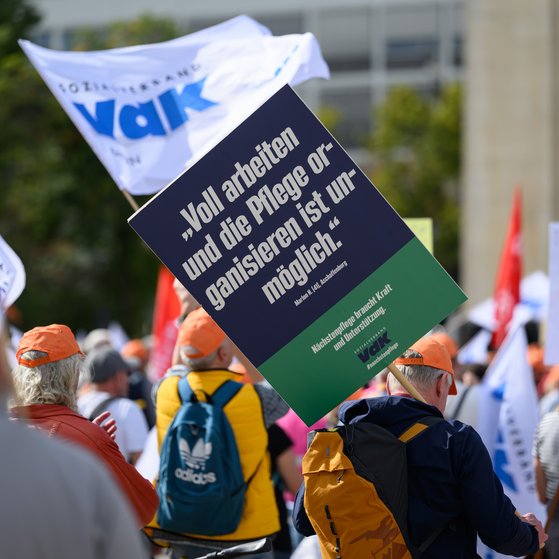 Menschen mit orangen M&uuml;tzen bei einer Demonstration, eine Person h&auml;lt ein Schild mit der Aufschrift 'Voll arbeiten und die Pflege organisieren ist unm&ouml;glich.'