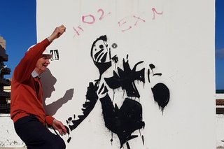 Auf dem Foto sieht man einen Mann vor einer Wand mit Graffiti stehen. Er hält einen Arm und ein Bein in die Luft. 