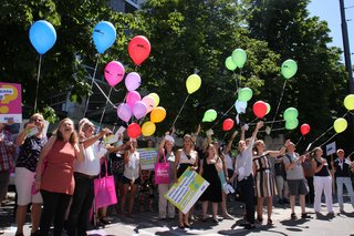 Auf dem Foto sieht man verschiedene Personen nebeneinander in einer Reihe stehen. Viele von ihnen halten bunte Luftballons in der Hand. 