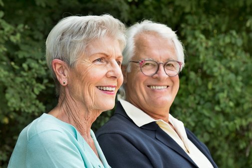 Das Foto zeigt ein &auml;lteres Ehepaar auf einer Parkbank in einem Park. Beide l&auml;cheln seitlich in die Kamera.