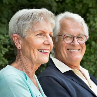 Das Foto zeigt ein älteres Ehepaar auf einer Parkbank in einem Park. Beide lächeln seitlich in die Kamera.