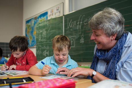 Eine Frau (rechts im Bild) sitzt in einem Klassenzimmer neben einem Jungen mit Down-Syndrom und hilft ihm bei einer Aufgabe.