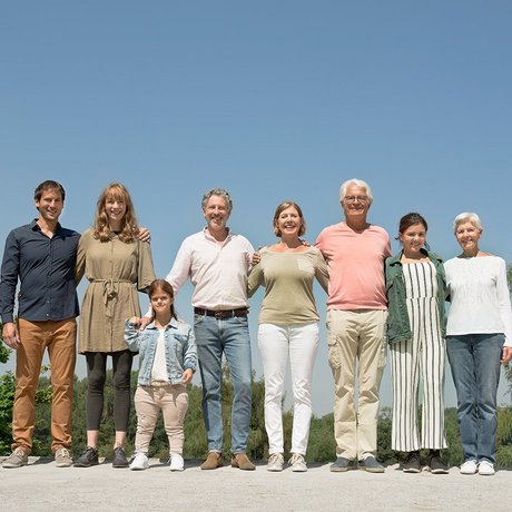 Das Bild zeigt eine Gruppe von älteren und jüngeren menschen mit und ohne Behinderung. Sie stehen auf einer Wiese und fassen sich an den Händen. Alle lächeln in die Kamera. 