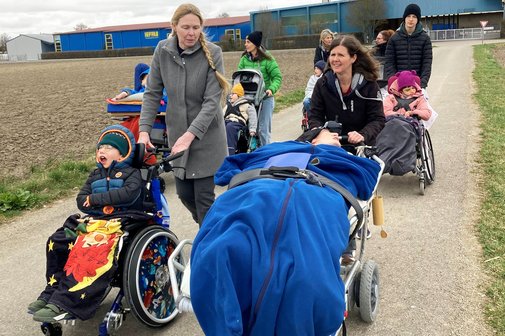 Die Szene zeigt eine Stra&szlig;e und daneben einen Acker, im Hintergrund ein blaues Industriegeb&auml;ude. Mehrere Frauen schieben die sieben pflegebed&uuml;rftigen Kinder und Jugendlichen in ihren Kinderw&auml;gen sowie einen Jugendlichen im Pflegebett. 