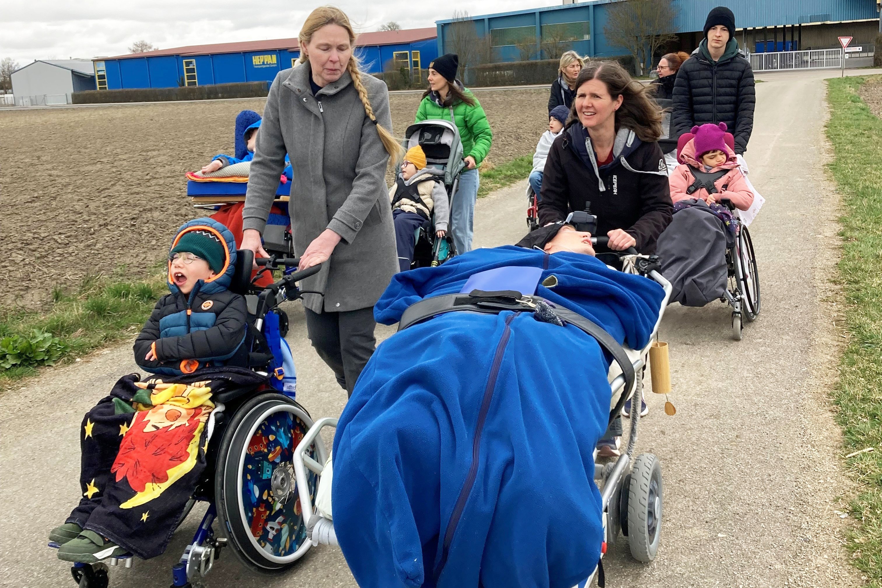 Die Szene zeigt eine Stra&szlig;e und daneben einen Acker, im Hintergrund ein blaues Industriegeb&auml;ude. Mehrere Frauen schieben die sieben pflegebed&uuml;rftigen Kinder und Jugendlichen in ihren Kinderw&auml;gen sowie einen Jugendlichen im Pflegebett. 
