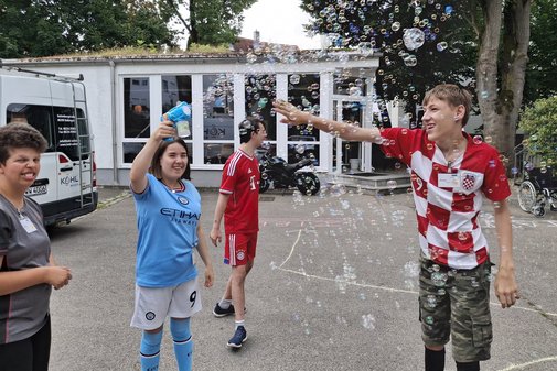 Vier Jugendliche, drei Jungen und ein M&auml;dchen, stehen vor einem Haus auf einem Parkplatz. Das M&auml;dchen spr&uuml;ht mit einer Seifenblasenmaschine Seifenblasen in die Luft. Einer der Jungen streckt seine Hand in die Menge der Seifenblasen. 