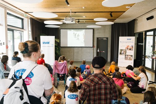 Kinder mit und ohne Behinderung sowie Freizeitbegleiterinnen sitzen auf dem Boden und sehen sich einen Film &uuml;ber eine VdK-Skifreizeit an.