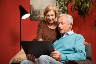 Das Bild zeitg ein älteres Paar in ihrem Wohnzimmer. Er sitzt in einem Sessel und hält ein Tablet in den Händen. Sie sitzt auf der Sessellehne und schaut mit ihm ins Tablet.
