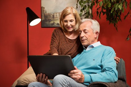 Das Bild zeitg ein älteres Paar in ihrem Wohnzimmer. Er sitzt in einem Sessel und hält ein Tablet in den Händen. Sie sitzt auf der Sessellehne und schaut mit ihm ins Tablet.