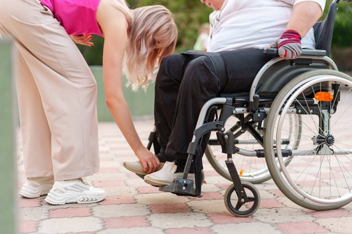 Eine junge Frau r&uuml;ckt einer &auml;lteren Person in einem Rollstuhl die F&uuml;&szlig;e auf den Fu&szlig;st&uuml;tzen zurecht. Young Carer &uuml;bernehmen schon fr&uuml;h Verantwortung f&uuml;r hilfs- und pflegebed&uuml;rftige Personen. 