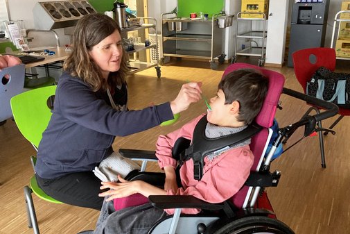 Eine Frau f&uuml;ttert mit einem L&ouml;ffel ein M&auml;dchen, das in einem Kinderwagen sitzt.