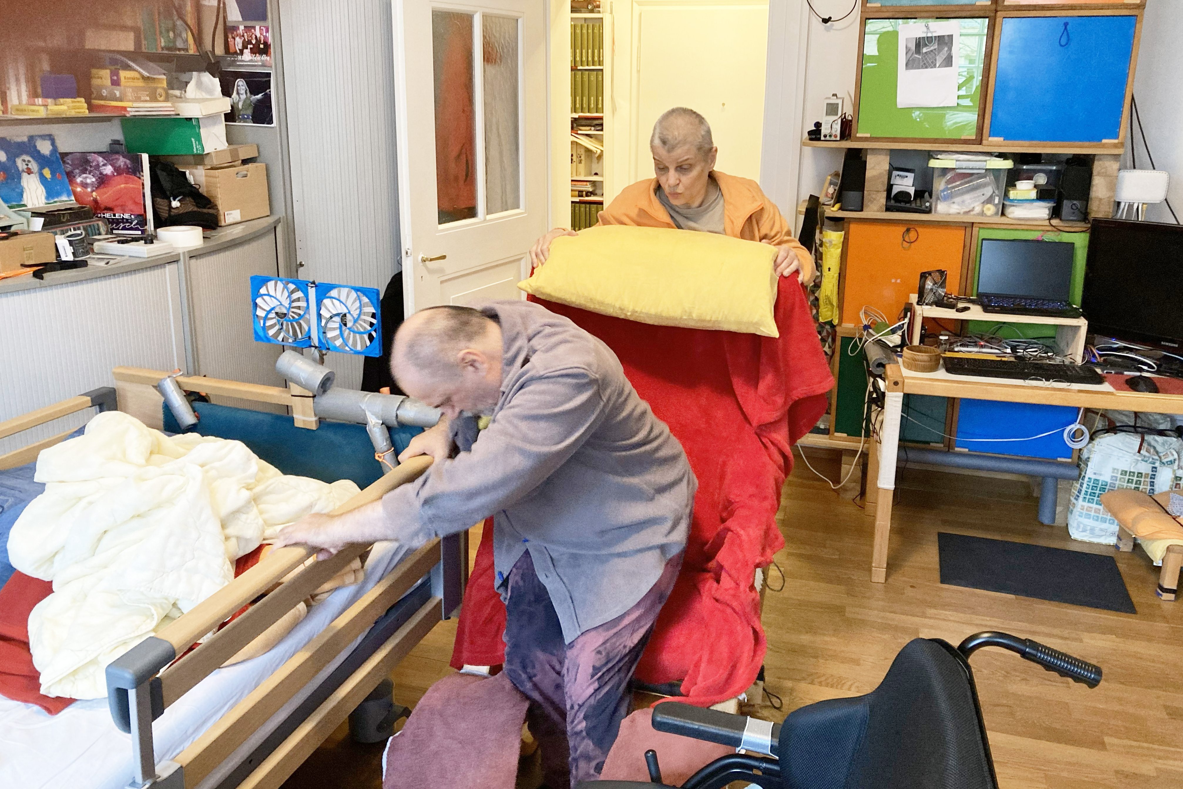 Holger Eichinger* (vorne) setzt sich vom Rollstuhl in seinen Sessel. Seine Frau Karin* hilft ihm dabei.