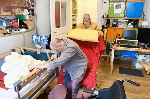 Holger Eichinger* (vorne) setzt sich vom Rollstuhl in seinen Sessel. Seine Frau Karin* hilft ihm dabei.