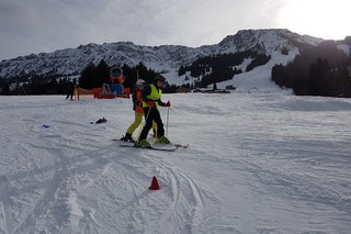 Das Foto zeigt einen Teilnehmenden am Monoskikurs auf der Piste beim Skifahren. Ein Skilehrer hält ihn zwischen seinen Beinen.
