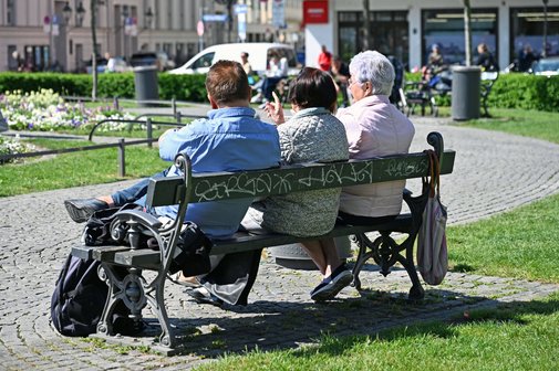 Zwei ältere Damen und ein älterer Herr sind von hinten fotografiert. Sie sitzen gemütlich auf einer Bank am Münchner Gärtnerplatz und unterhalten sich. 
