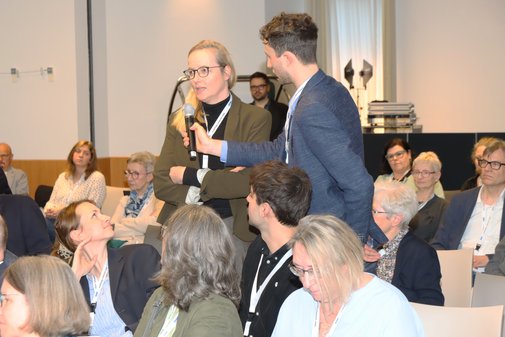 Ein Mann (rechts) steht neben einer Frau und hält ihr ein Mikro vor das Gesicht.