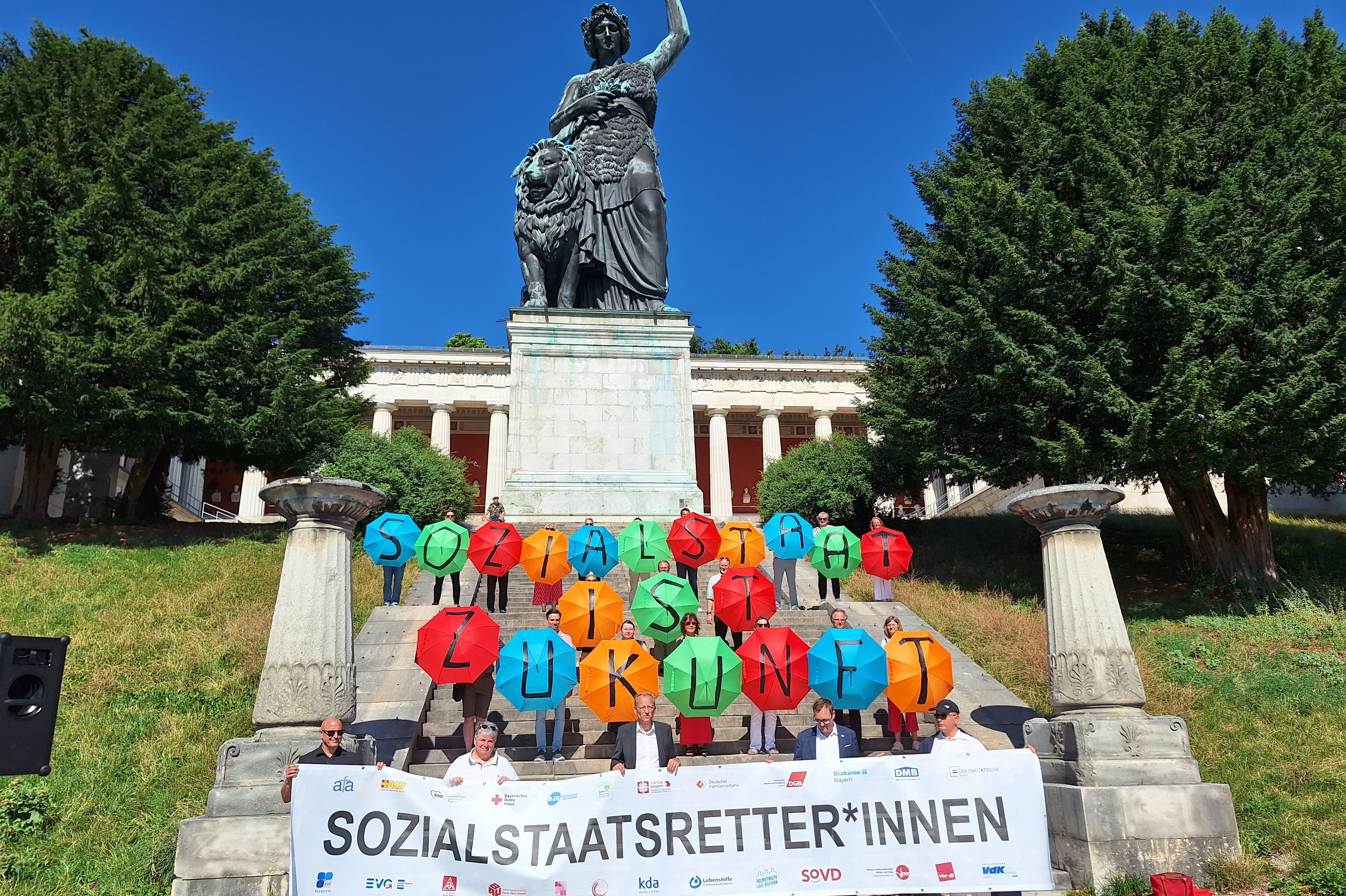Teilnehmende des Aktionsbündnisses „Gemeinsam für einen starken und zukunftsfähigen Sozialstaat“ stehen in München vor der Bavaria in mehreren Reihen und halten ein gemeinsames Banner.