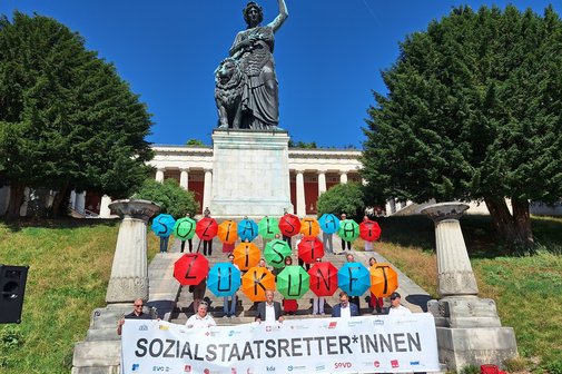 Teilnehmende des Aktionsbündnisses „Gemeinsam für einen starken und zukunftsfähigen Sozialstaat“ stehen in München vor der Bavaria in mehreren Reihen und halten ein gemeinsames Banner.