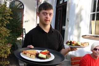 Auf dem Foto sieht man Wolfgang Oefner. Er arbeitet im Café Vielfalt in Regensburg. Er hat zwei Teller mit Essen in der Hand. Im Hintergrund sieht man den Garten des Cafés.
