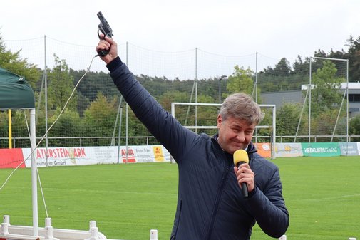 Marcus Mittermeier steht am Rand einer Leichtathletik-Laufbahn im Freien. Er hat eine Pistole in der Hand, mit der er in die Luft zielt. Mit einer Platzpatrone gibt er den Startschuss für einen Spendenlauf bei einer Inklusionsveranstaltung in Niederbayern.
