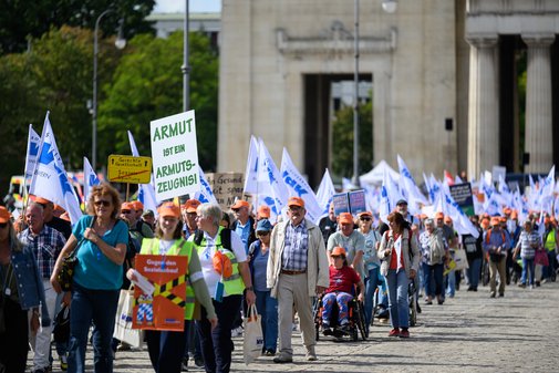 Demonstrationszug des VdK Bayern mit vielen Teilnehmenden, die Fahnen und Schilder gegen Sozialabbau tragen, einige mit orangen M&uuml;tzen, vor einem gro&szlig;en steinernen Torbogen