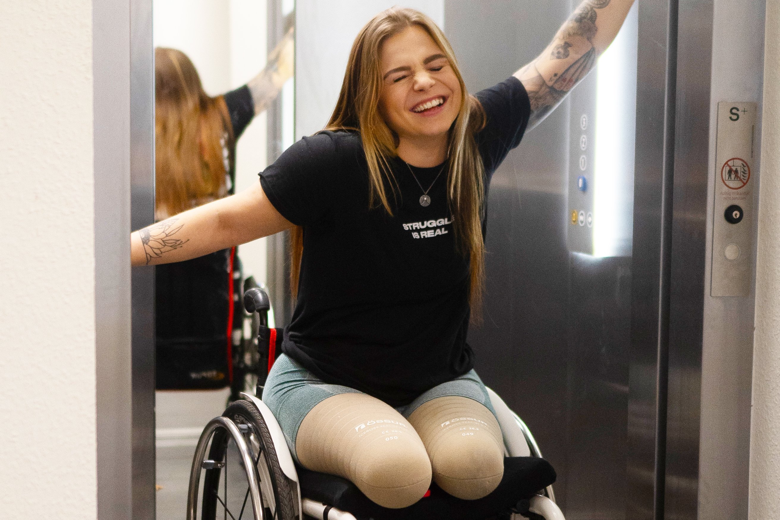 Das Foto zeigt Angie Berbeuer in einem Aufzug. Sie sitzt in einem Rollstuhl und hält mit beiden Armen den Aufzug offen. Ihre Augen sind geschlossen und sie lacht. 
