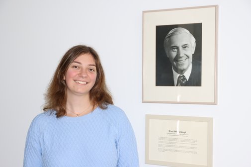 Eine junge Frau steht an einer weißen Wand, an der ein Schwarz-Weiß-Foto eines älteren Mannes hängt. Sie lächelt. Das Bild zeigt Karoline Weishäupl neben dem Bild ihres Großvaters Karl Weishäupl, der den VdK zusammen mit anderen 1946 ins Leben gerufen hat.