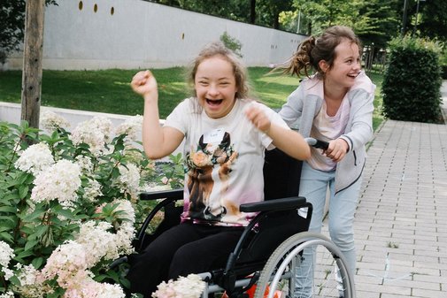 Ein M&auml;dchen mit Downsyndrom wird von einem anderen M&auml;dchen im Rollstuhl geschoben. Sie kommen etwas vom Weg ab und ein Rad des Rollstuhls steht im Blumenbeet. Beide lachen.