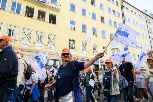 Menschen mit orangen M&uuml;tzen und Fahnen mit der Aufschrift 'VdK Bayern' bei einer Demonstration vor einem gelben Wohnhaus mit ge&ouml;ffneten Fenstern, aus denen weitere Menschen zuschauen