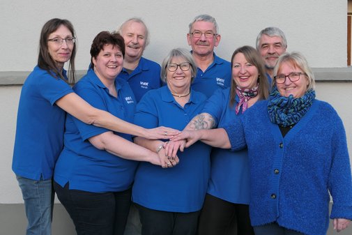 Acht VdK-Ehrenamtliche stehen eng nebeneinander und legen ihre H&auml;nde &uuml;bereinander. Sie tragen blaue VdK-T-Shirts und lachen in die Kamera. 