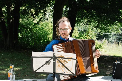 Der Musiker Simon Japha sitzt mit seinem Akkordeon vor einem Baum und spielt.