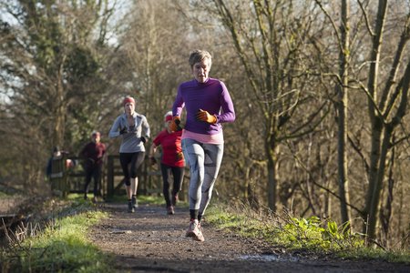 Eine ältere Frau joggt auf einem Feldweg, hinter ihr sind weitere Jogger zu sehen. Das Joggen in der Gruppe macht vielen Menschen mehr Spaß als allein. Um Gewohnheiten zu brechen und gute Vorsätze, wie mehr Sport zu machen, umzusetzen, kann man sich einer Gruppe anschließen.