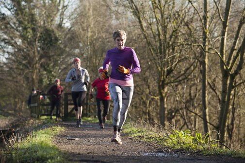 Eine ältere Frau joggt auf einem Feldweg, hinter ihr sind weitere Jogger zu sehen. Das Joggen in der Gruppe macht vielen Menschen mehr Spaß als allein. Um Gewohnheiten zu brechen und gute Vorsätze, wie mehr Sport zu machen, umzusetzen, kann man sich einer Gruppe anschließen.
