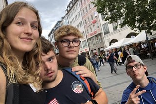 Das Foto zeigt drei Teilnehmer der inklusiven Ferienfreizeit und eine Freizeitbegleiterin in der Münchner Innenstad. Sie hält einen Teilnehmer im Arm. 