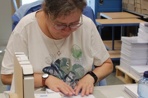 Eine Mitarbeiterin von Dimetria sitzt an ihrem Arbeitsplatz und faltet einen Brief. Vor ihr liegen VdK-Brosch&uuml;ren und Briefumschl&auml;ge.