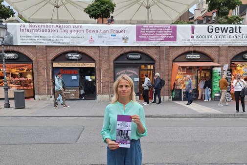 VdK-Landesvorsitzende Verena Bentele steht vor dem aufgeh&auml;ngten Banner auf dem Viktualienmarkt in M&uuml;nchen und h&auml;lt einen Flyer zur Aktion in ihren H&auml;nden.
