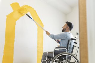 Ein Mann in einem Rollstuhl malt die Wand seiner Wohnung mit einem Farbroller gelb an. 