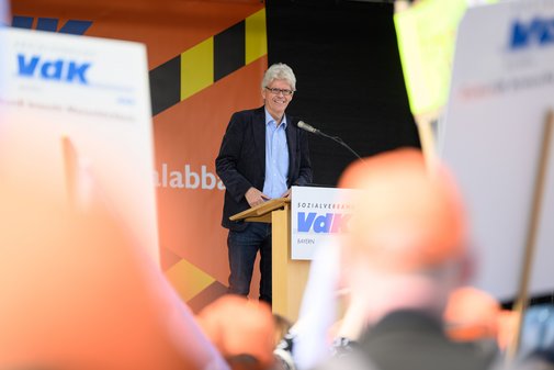 VdK-Landesgesch&auml;ftsf&uuml;hrer Michael Pausder steht l&auml;chelnd hinter einem Rednerpult mit dem Schriftzug 'Sozialverband VdK Bayern' und spricht in ein Mikrofon, im Vordergrund unscharfe erhobene H&auml;nde und Schilder.