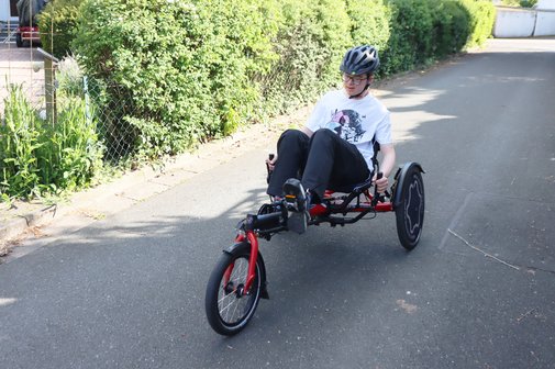 Der Jugendliche Balthasar Bars sitzt auf einem Liegerad und fährt an einem sonnigen Tag einen Gehweg entlang, an dessen Saum ein Zaun mit grüner Hecke zu sehen ist.