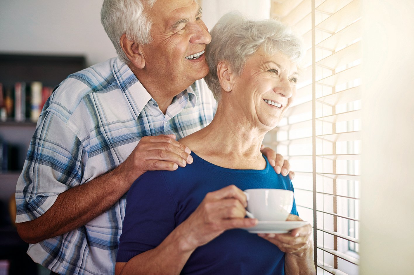 Auf dem Foto sieht man zwei &auml;ltere Menschen, die am Fenster stehen und nach drau&szlig;en schauen. 