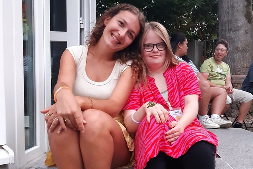 Zwei M&auml;dchen sitzen einer Treppe. Sie lehnen ihre K&ouml;pfe aneinander und l&auml;cheln in die Kamera. Im Hintergrund sitzen zwei Personen auf St&uuml;hlen und unterhalten sich. 