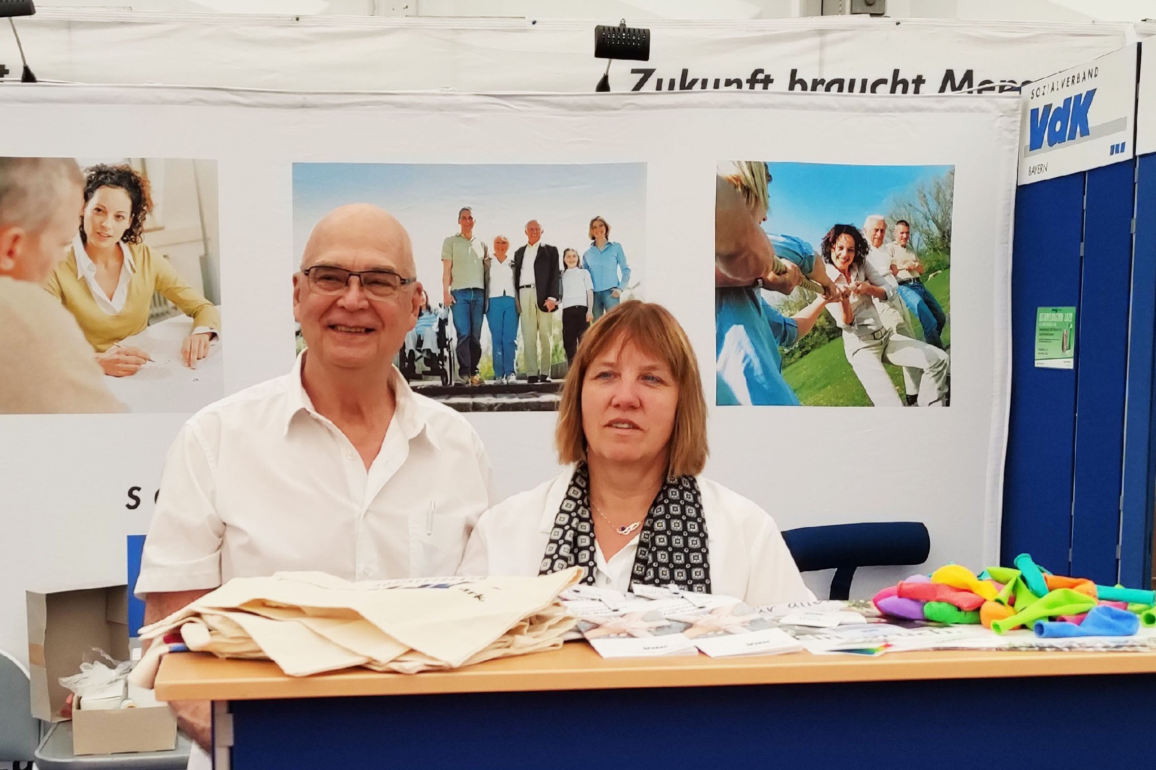 Horst Gottschild (links) und Kerstin Seinige stehen am Tresen eines VdK-Stands auf einer Veranstaltung, vor ihnen liegen Give-Aways wie Luftballons und Stofftaschen, im Hintergrund sind VdK-Plakate zu sehen.