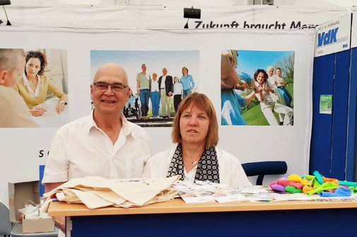 Horst Gottschild (links) und Kerstin Seinige stehen am Tresen eines VdK-Stands auf einer Veranstaltung, vor ihnen liegen Give-Aways wie Luftballons und Stofftaschen, im Hintergrund sind VdK-Plakate zu sehen.