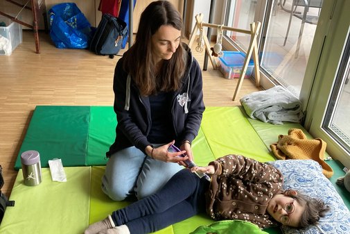 Ein etwa zehn Jahre altes M&auml;dchen liegt auf mehreren gr&uuml;nen, weichen Bodenmatten. Unter der braunen Fleecejacke schaut ein Schlauch heraus. Es ist eine Magensonde. Neben ihr hockt eine Pflegerin und spritzt Nahrung in den Schlauch der Sonde.