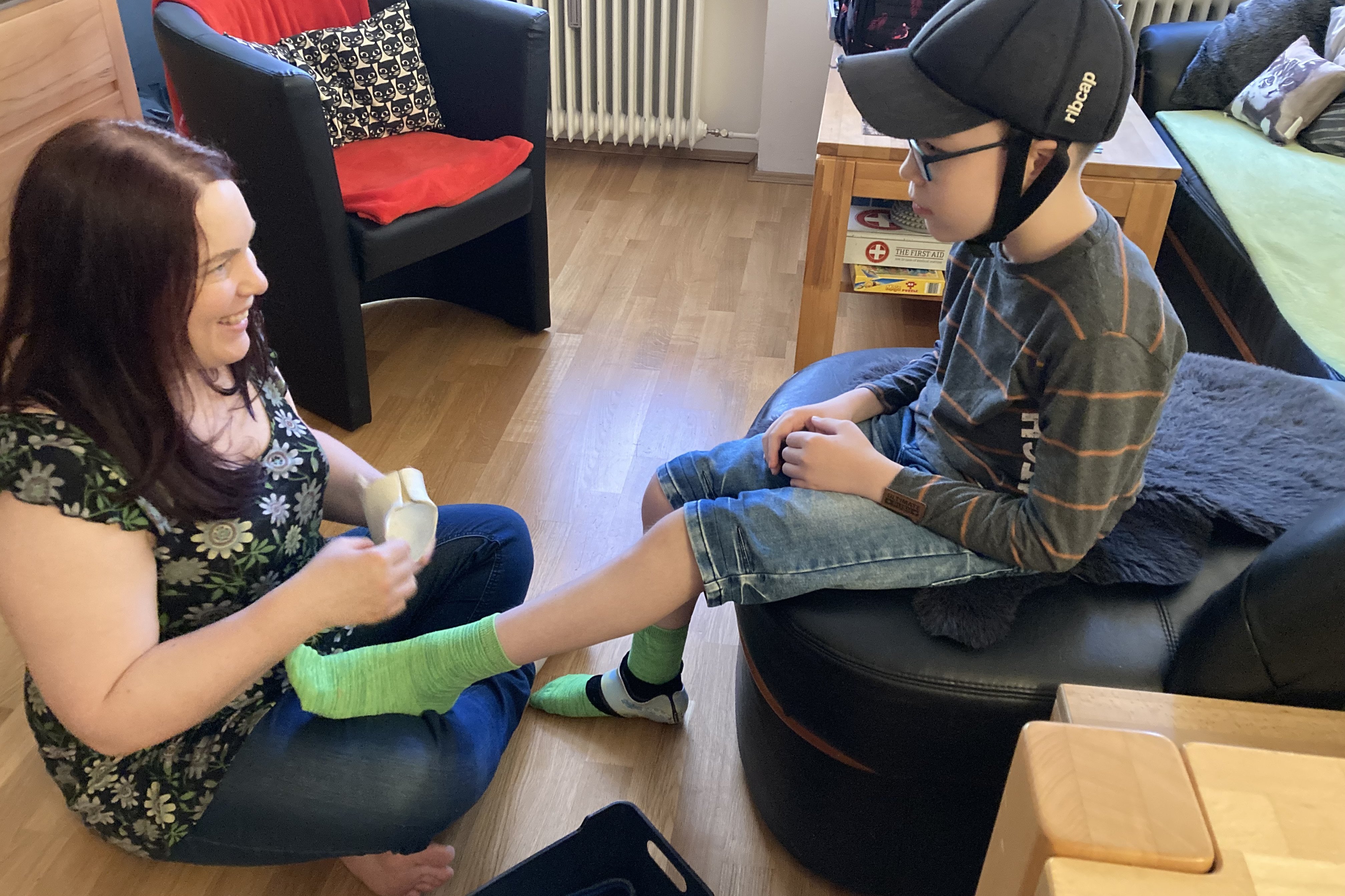 Auf dem Foto sieht man Jana mit ihrem jüngsten Sohn im Wohnzimmer. Sie sitzt auf dem Boden und hilft ihm, die Socken anzuziehen. 