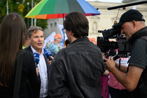 Mann im Anzug wird von zwei Interviewern mit Mikrofonen und einem Kameramann mit gro&szlig;er Kamera vor einer Menschenmenge und einem bunten Regenschirm gefilmt